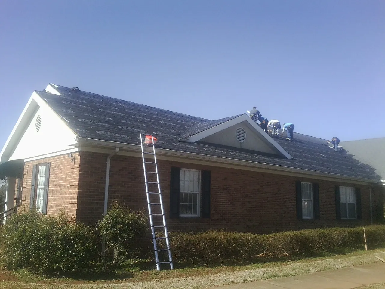 Crew tearing shingles off a brick ranch roof with a ladder set — Emergency Roof Repair work by our crew in Cedar Lake