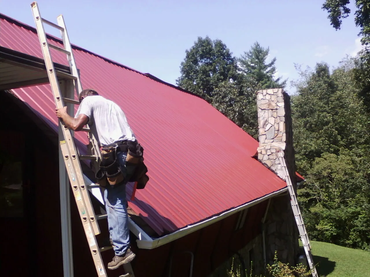 Roofer on a ladder beside a red metal roof and stone chimney — Soffit & Fascia Repair by Roofing Brothers in Cedar Lake
